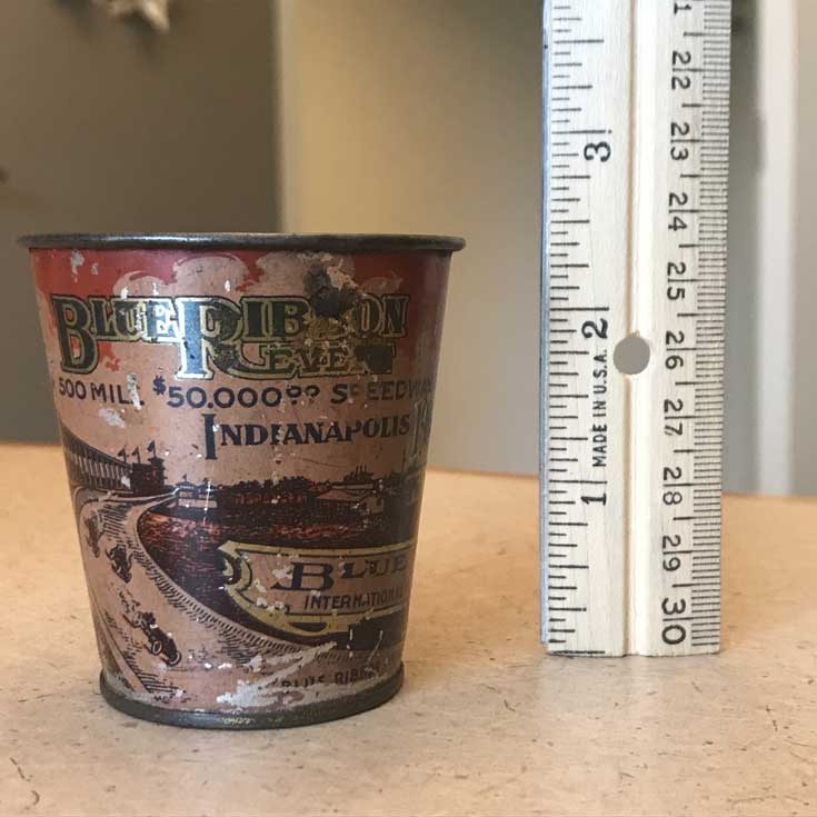 Metal cup with Blue Ribbon written across top and an illustration of the racetrack with cars on it that goes around the cup. It is less than 3 inches tall as shown by a ruler held up next to the cup. 