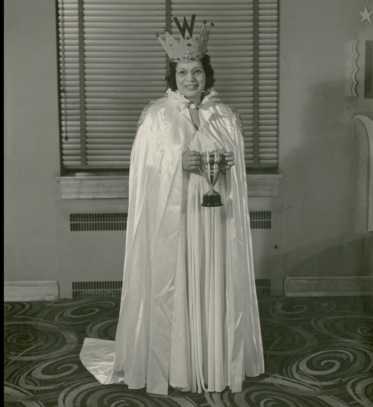 Woman in gown and cloak wearing a crown with a W and holding a trophy.