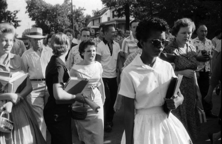 Black woman, Elizabeth Eckford, in white dress holding a book while white people surround her. 