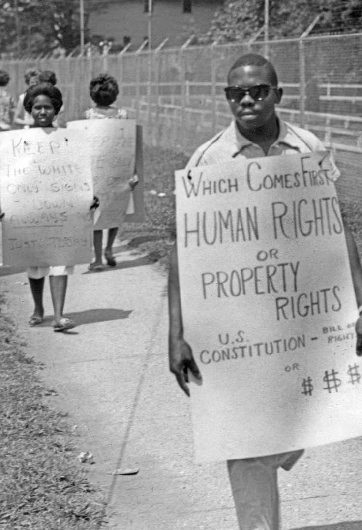 Black man in sunglasses walking on sidewalk with holding sign that says "Which comes first human rights or property rights"