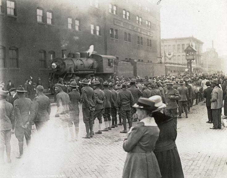 A crowd of people, including African American soldiers, watch as a train transporting soldiers comes to a stop. Trains arrived throughout the day, bringing troops to participate in the Welcome Home Day Parade and festivities.