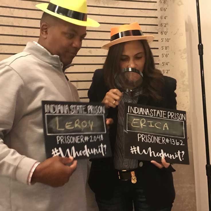 Two Whodunit attendees pose in front of the old fashioned mug shot screen wearing hats and holding chalkboard signs that have writing on them.