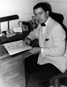 Jim Jones is seated at a desk holding a three-ring notebook. He is wearing a light jacket and bow tie. The picture is dated December 8, 1956.